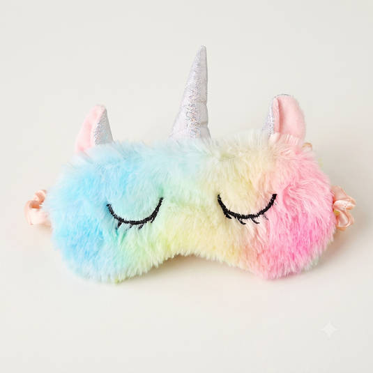 A plush unicorn sleep mask with a pastel rainbow gradient, featuring embroidered closed eyes, a silver glitter horn, and pink ears, shown on a plain light background.