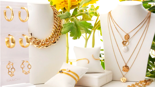 Gold jewelry including necklaces, bracelets, and earrings displayed on mannequin busts and stands with a white box and green plants in the background.